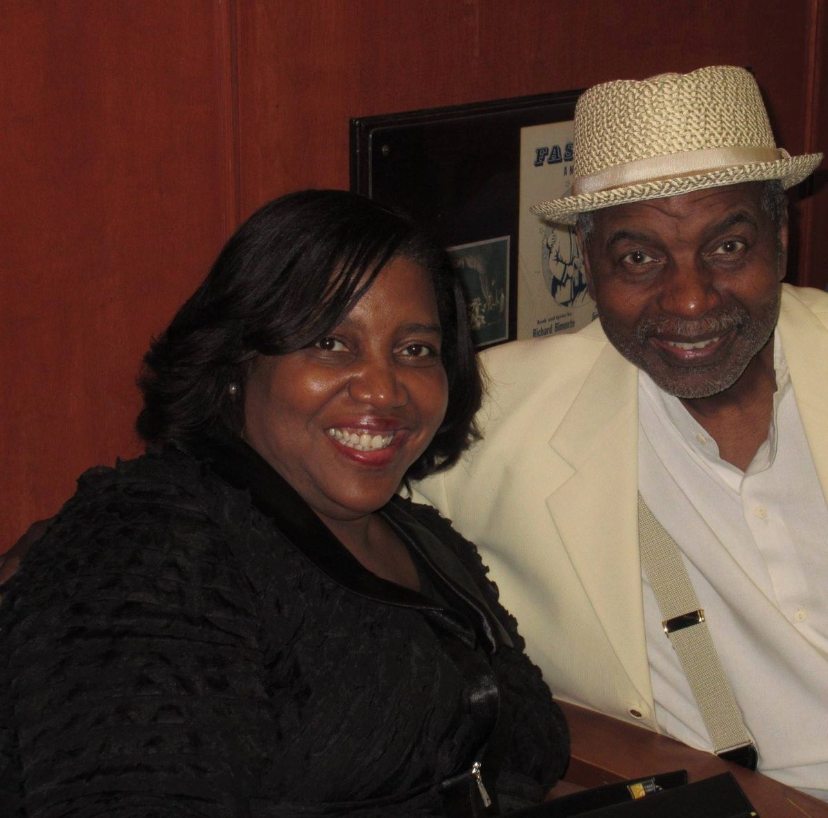 Dr. Evelyn Collins smiles beside Richard Wesley, who wears a cream jacket and straw hat, in front of a wood-paneled wall.
