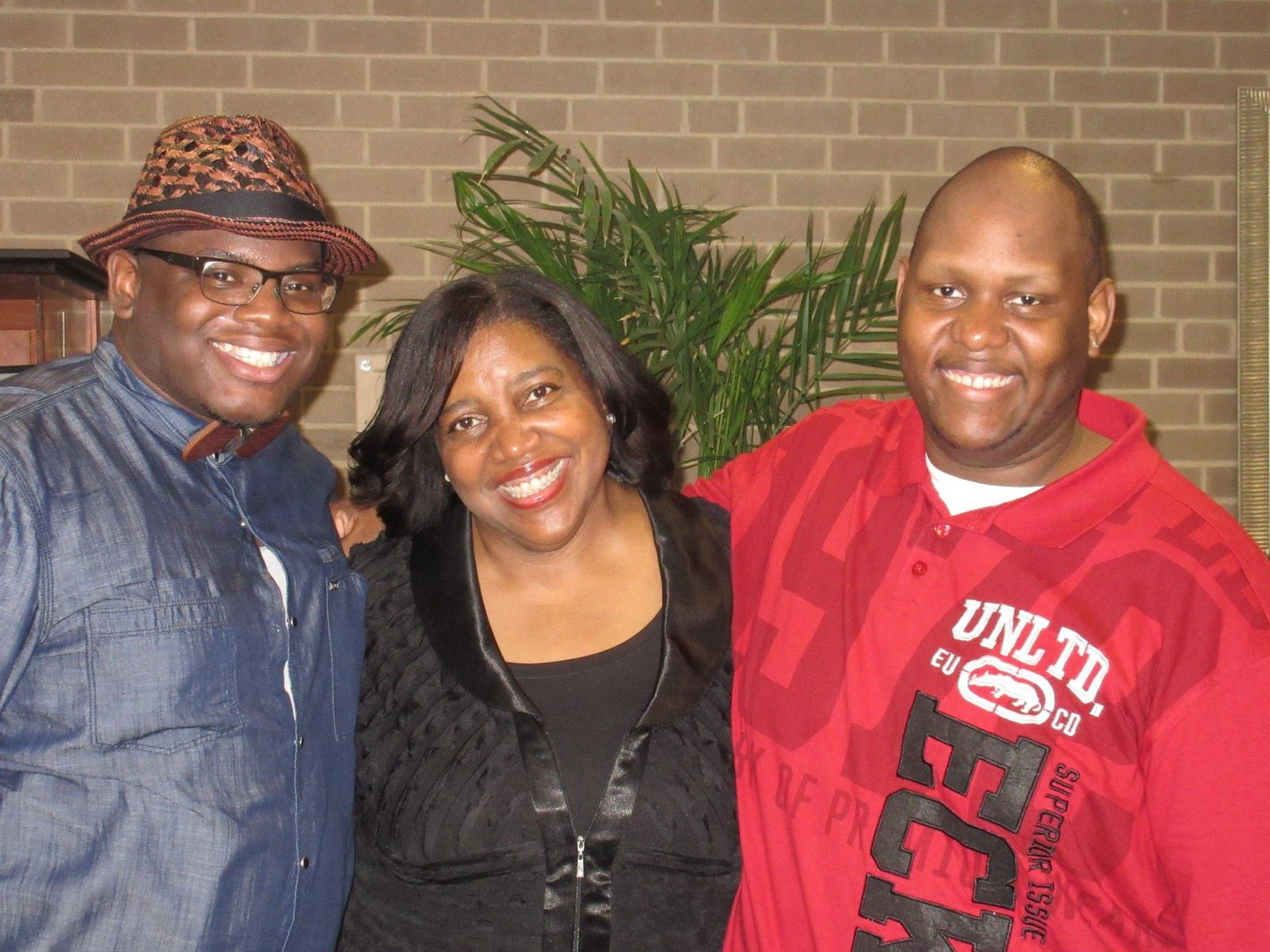 Dr. Evelyn Collins smiles between two young men, one in a straw fedora, glasses and wooden bow tie, the other in a red shirt, in front of a brick wall and a potted palm.