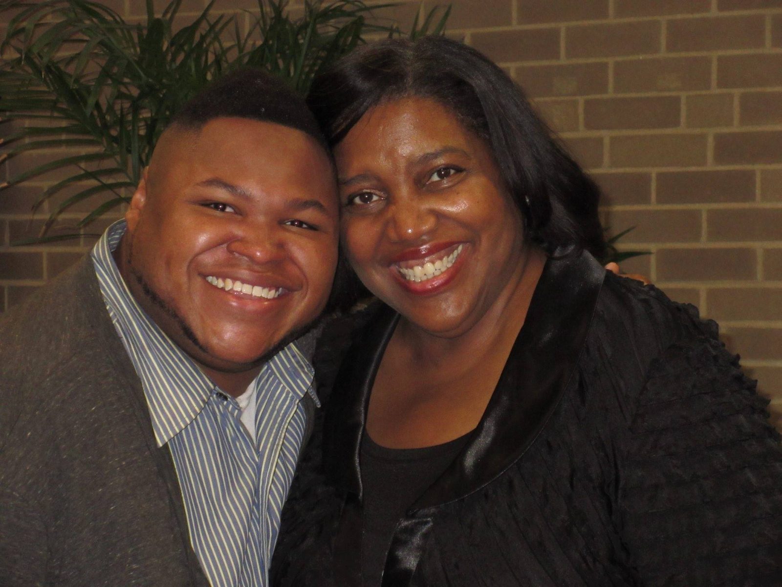 Dr. Evelyn Collins and a young man in a striped shirt and gray cardigan pose cheek to cheek in front of a brick wall.