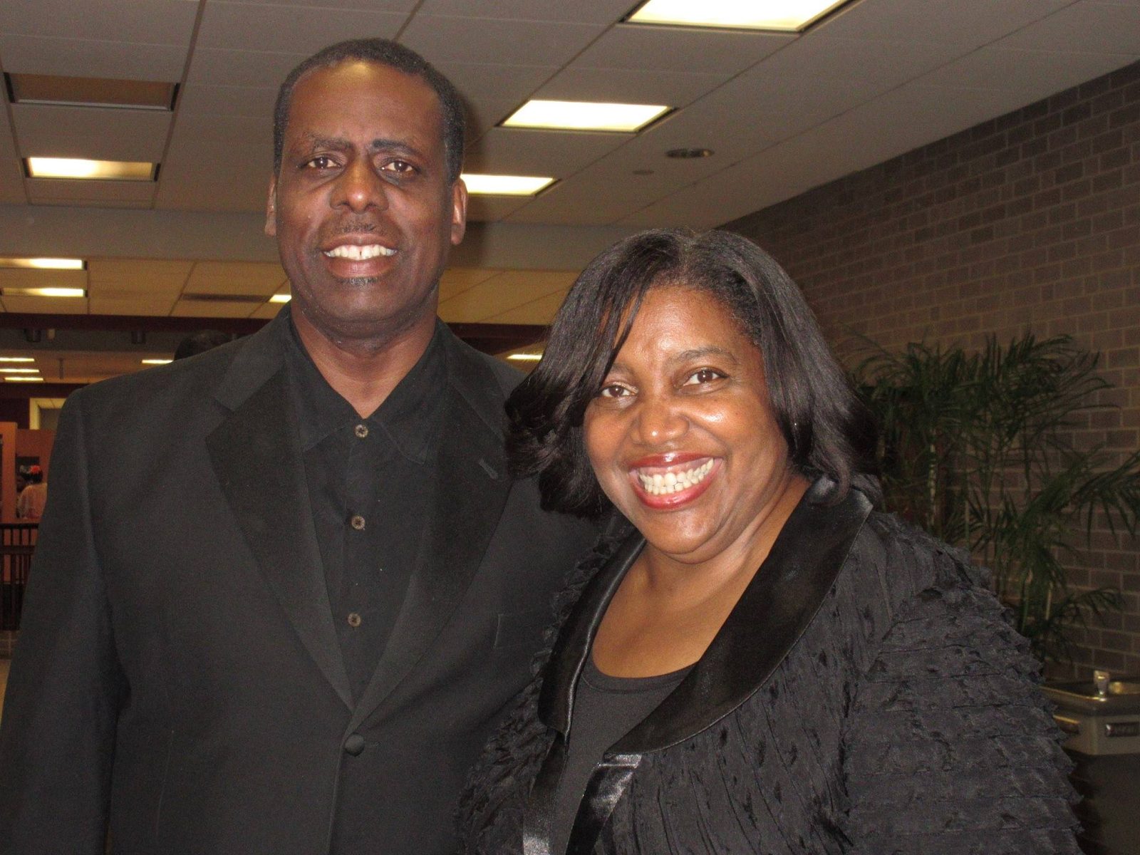 Dr. Evelyn Collins stands beside a tall man in a black suit in a brightly lit lobby with a brick wall and potted palms.