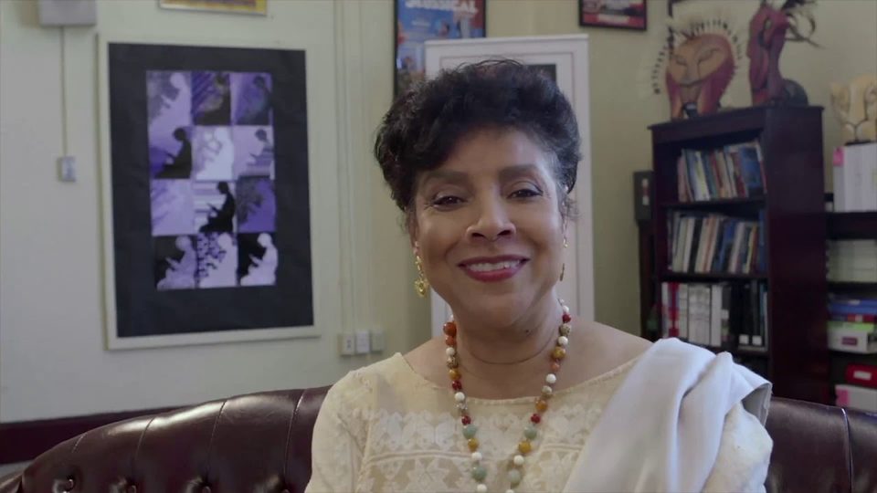 A woman in a cream blouse and a beaded necklace smiles from a leather couch in an office lined with books and theatre posters, in a still from In Celebration of the Arts 2019.