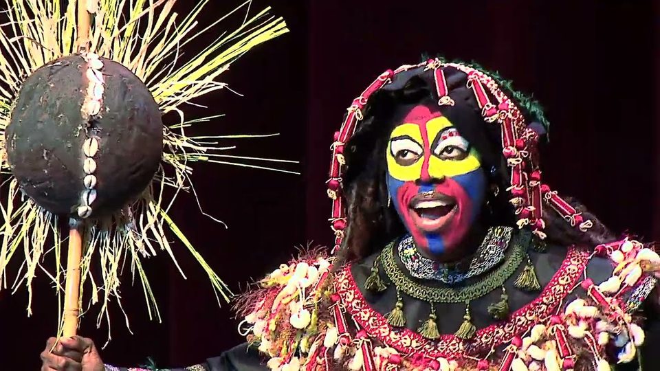 A performer in bright face paint and a beaded headdress holds up a gourd staff on stage in The Lion King, Jr.