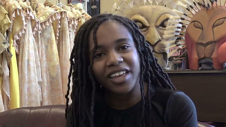 A student speaks from a leather couch in front of costume racks and lion and bird masks, in a still from the Excellence in Theatre Education video.