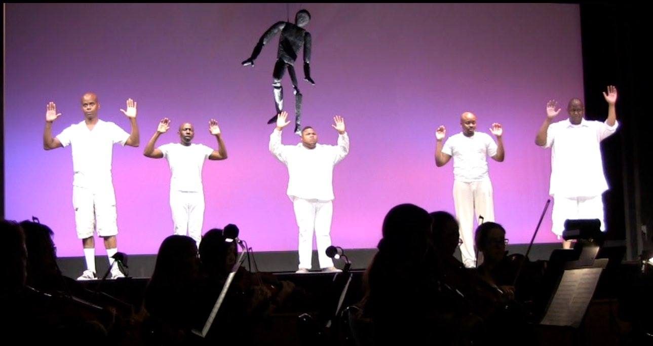 Five performers dressed in white raise their hands on a magenta-lit stage in The Central Park Five, an opera directed by Dr. Evelyn Collins.
