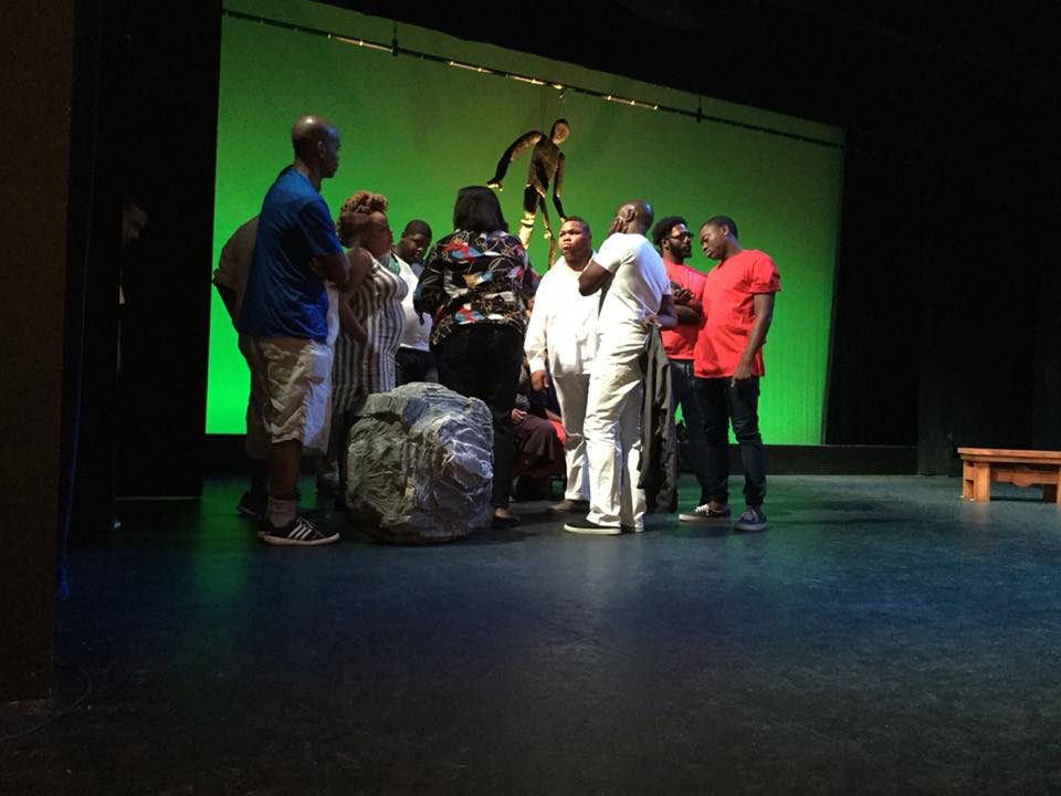 Members of the company gather in a close circle on a dim stage before a green-lit backdrop, beside a large rock set piece.