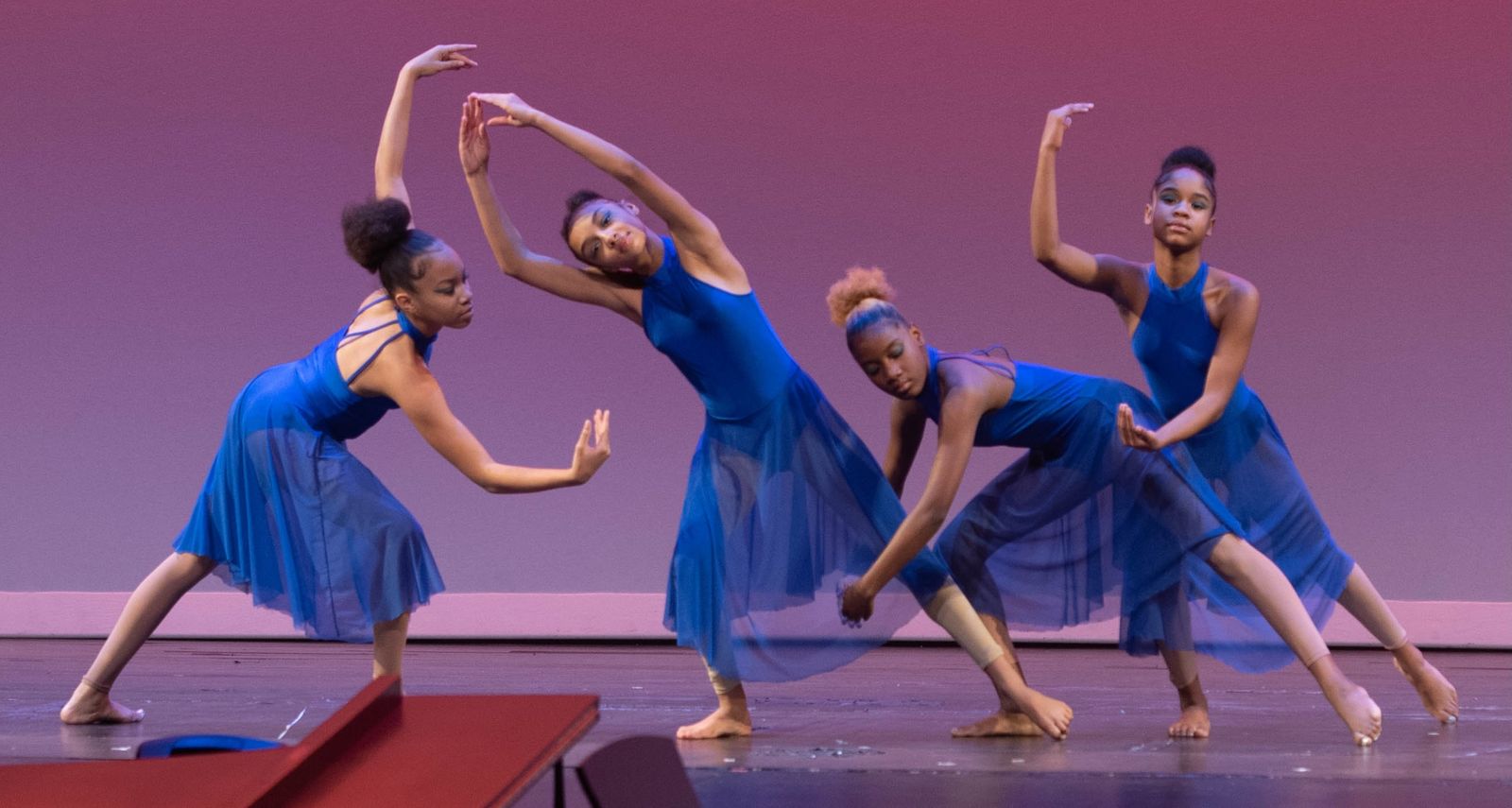 Four young dancers in blue dresses strike angular poses in front of a rose-colored backdrop.