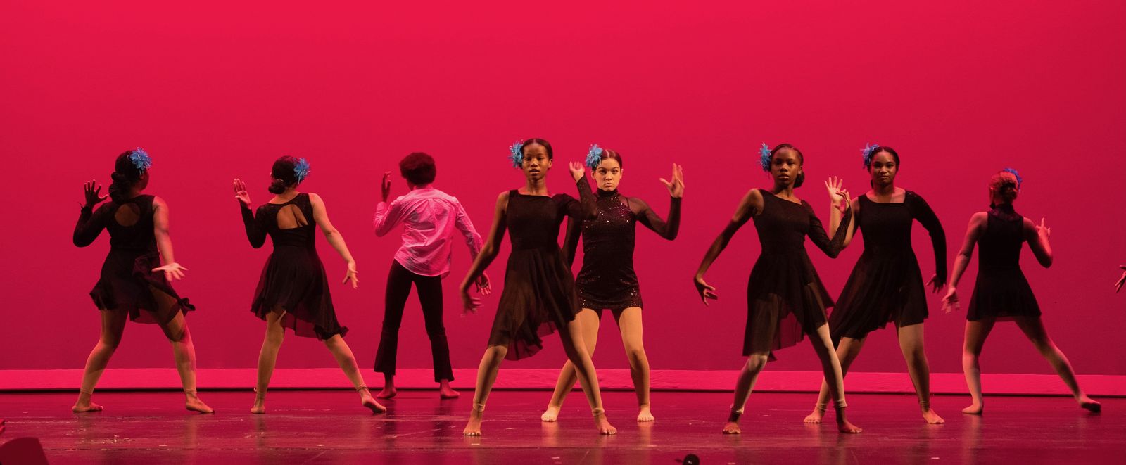 Dancers in black dresses with blue hair flowers pose mid-routine on a stage washed in red light.