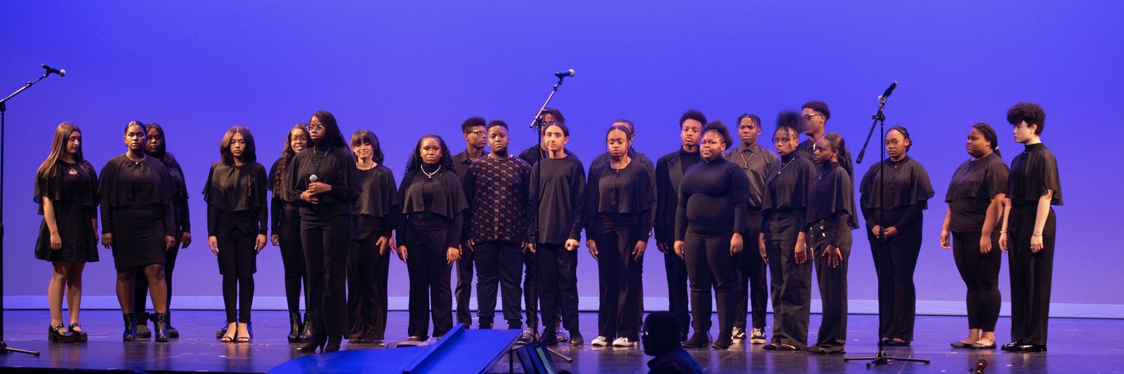 A large company of young performers dressed in black stands in a line across the stage between microphone stands.