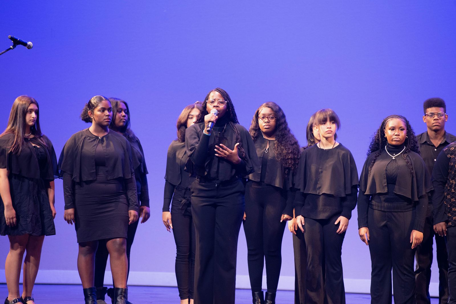 A young vocalist sings into a handheld microphone, one hand raised, with an ensemble in black behind her.