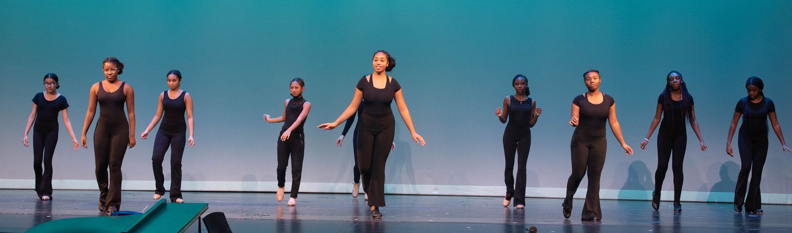 A line of young dancers in black rehearsal wear steps in unison before a teal backdrop.