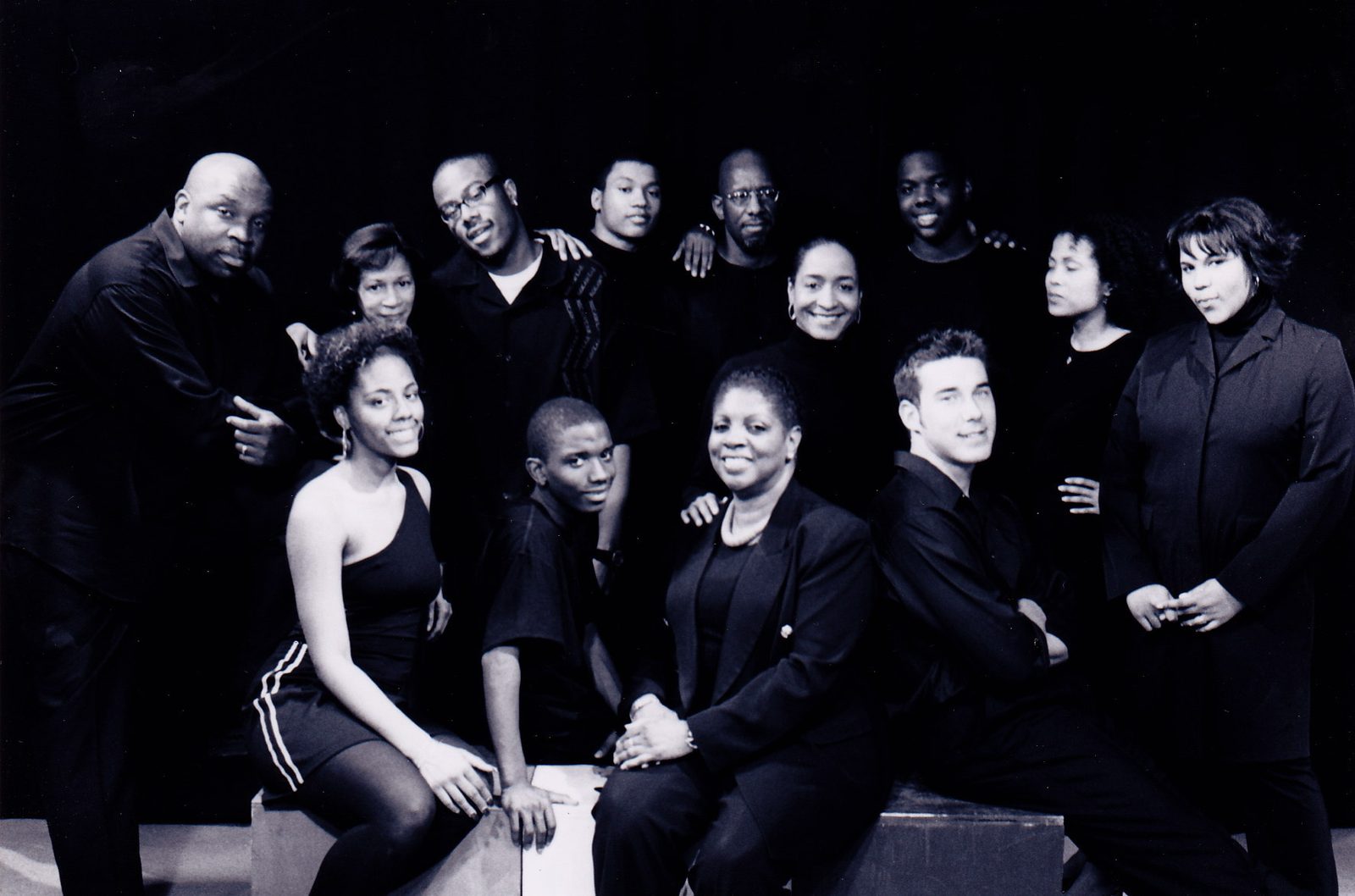 Black-and-white group portrait of thirteen company members dressed in black, posed around wooden boxes against a dark backdrop.