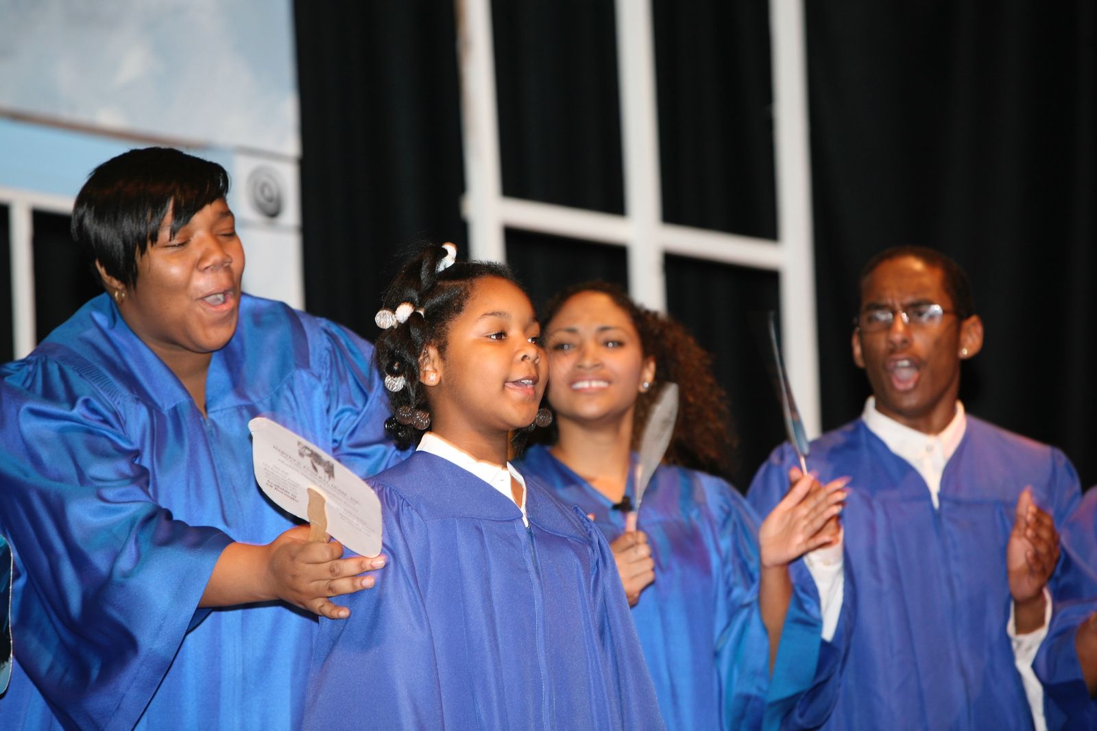 Singers in royal blue choir robes perform with hand fans in front of a set piece of tall window frames.